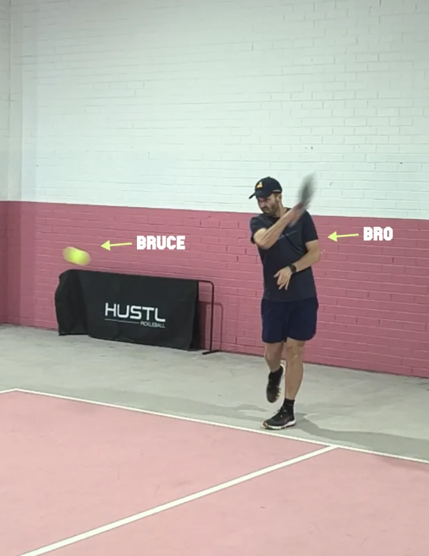 A pickleball player hitting a powerful serve with the BIOBall "Bruce" on an indoor pink court, with a Hustl Pickleball rebounder in the background
