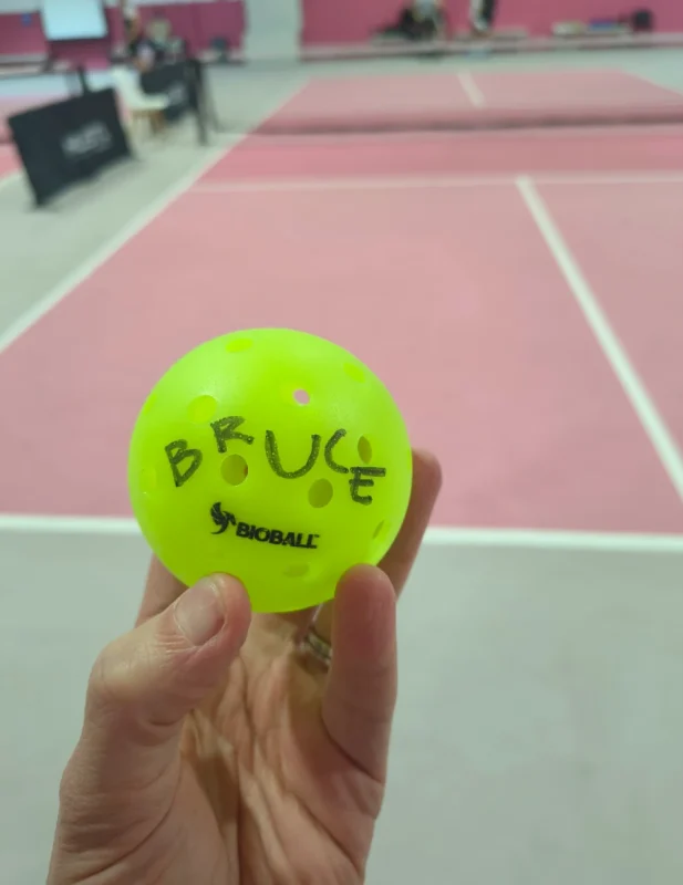 A hand holding a green BIOBall pickleball with the name "Bruce" written on it, in front of a pink indoor pickleball court for the bioball pickleball review